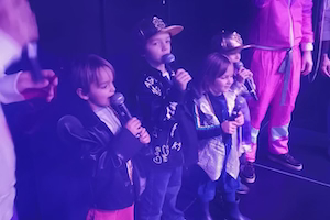 trois enfants âgés de 4 à 8 ans chantent sur la scène immersive, un micro à la main.